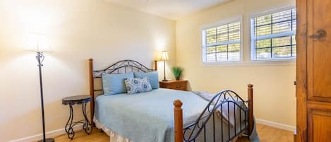 Un dormitorio con cama, mesita de noche, lámpara, ventilador de techo y ventana con persianas.