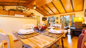 A dining area with a wooden table set for two, surrounded by chairs, and a view of the outdoors through large windows.