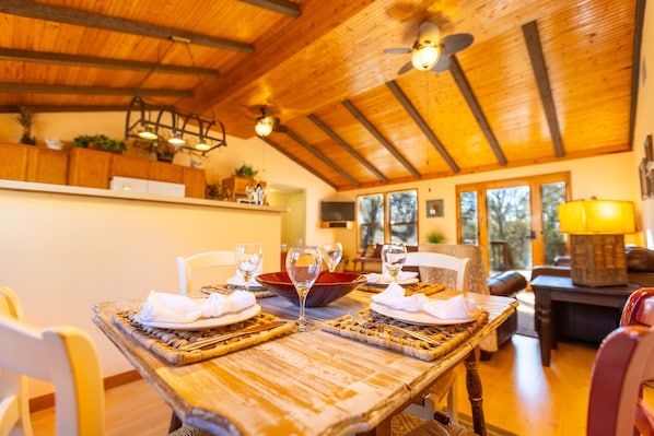 A dining area with a wooden table set for two, surrounded by chairs, and a red bowl centerpiece. - Wimberley Getaway with Private Pool and Fire Pit (Wimberley)