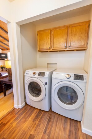 A laundry room with two Samsung front-loading washing machines, wooden cabinets above, and a wooden floor. - Wimberley Getaway with Private Pool and Fire Pit (Wimberley)