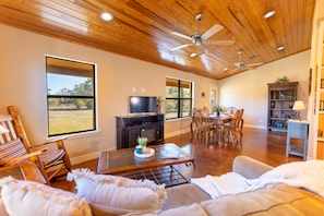 A spacious living room with a wooden ceiling, large windows, a television, a dining area, and a bookshelf. - Star Ranch Cottage- Starry Nights & Cozy Comfort (Wimberley)