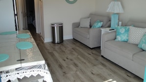 Un salon chaleureux avec un canapé, une table basse et une lampe. On y trouve une poubelle et une pancarte sur laquelle est écrit : « Bienvenue à Beach House ».