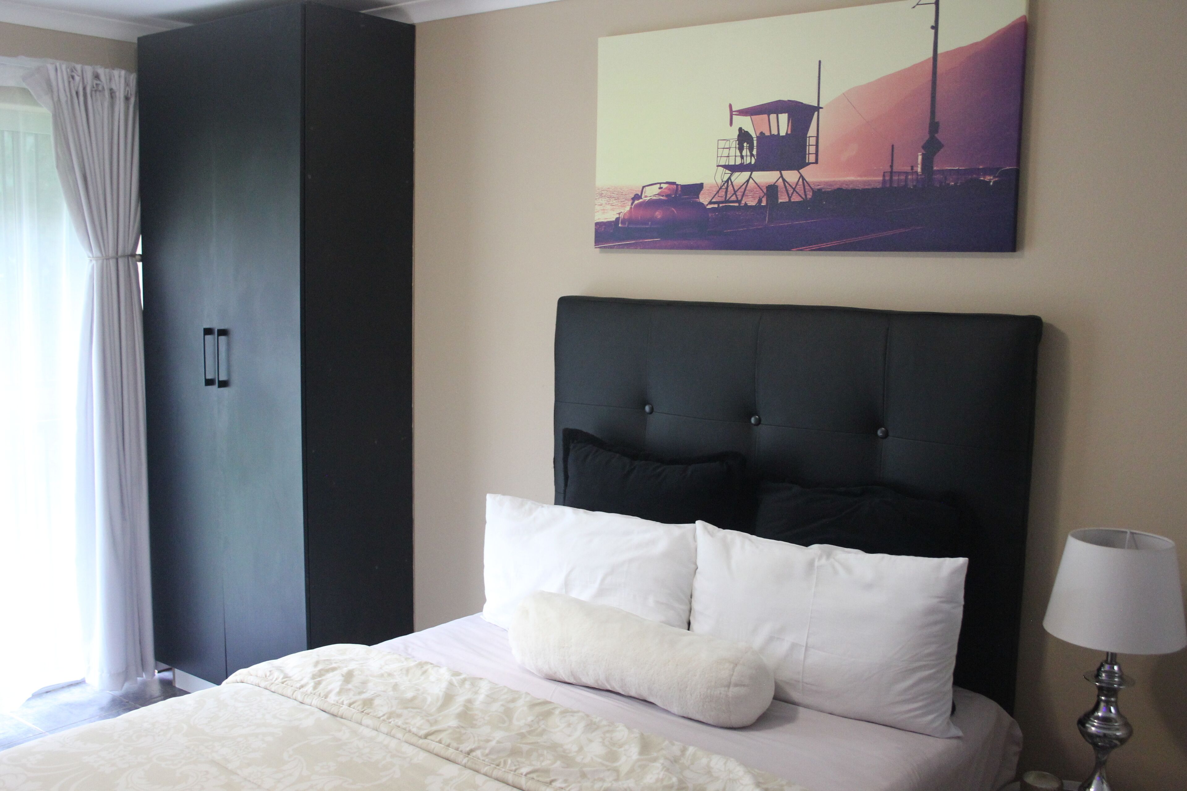 A neatly made bed with white bedding and two pillows, a dark headboard, a wall-mounted artwork depicting a coastal scene, and a tall wardrobe.