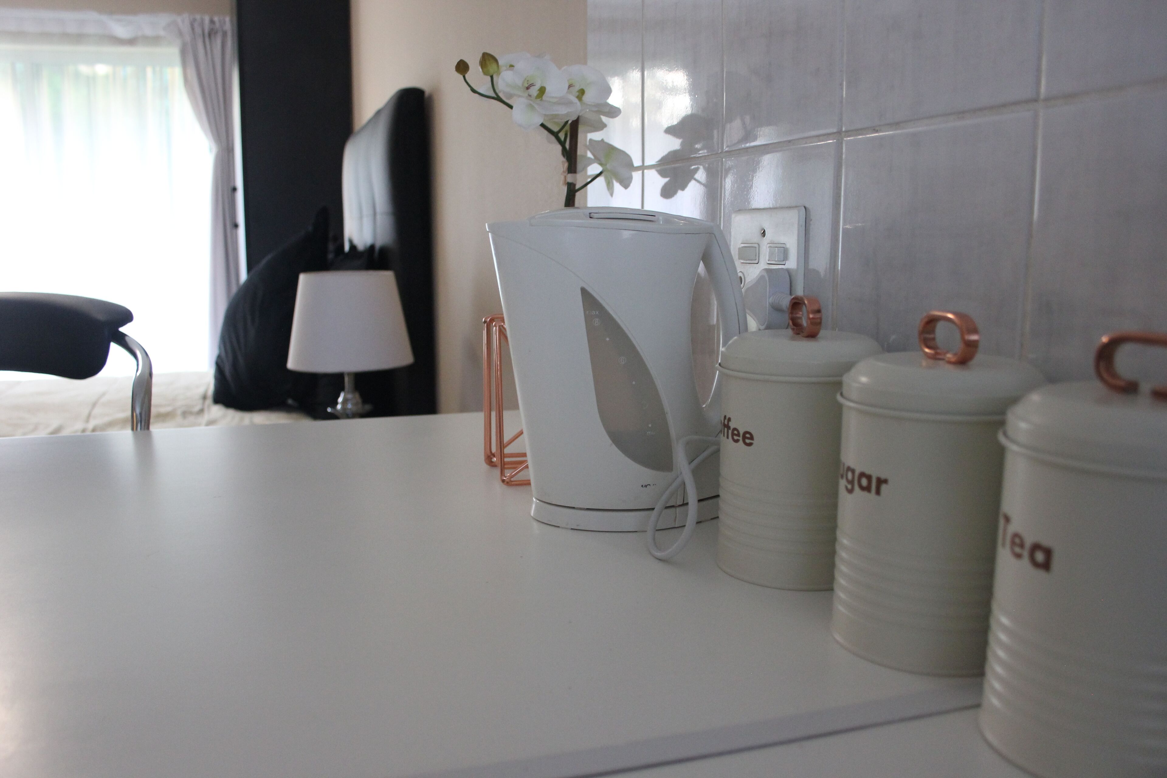 Una cafetera blanca, recipients de sucre i te, una làmpada blanca i una flor sobre una superfície.