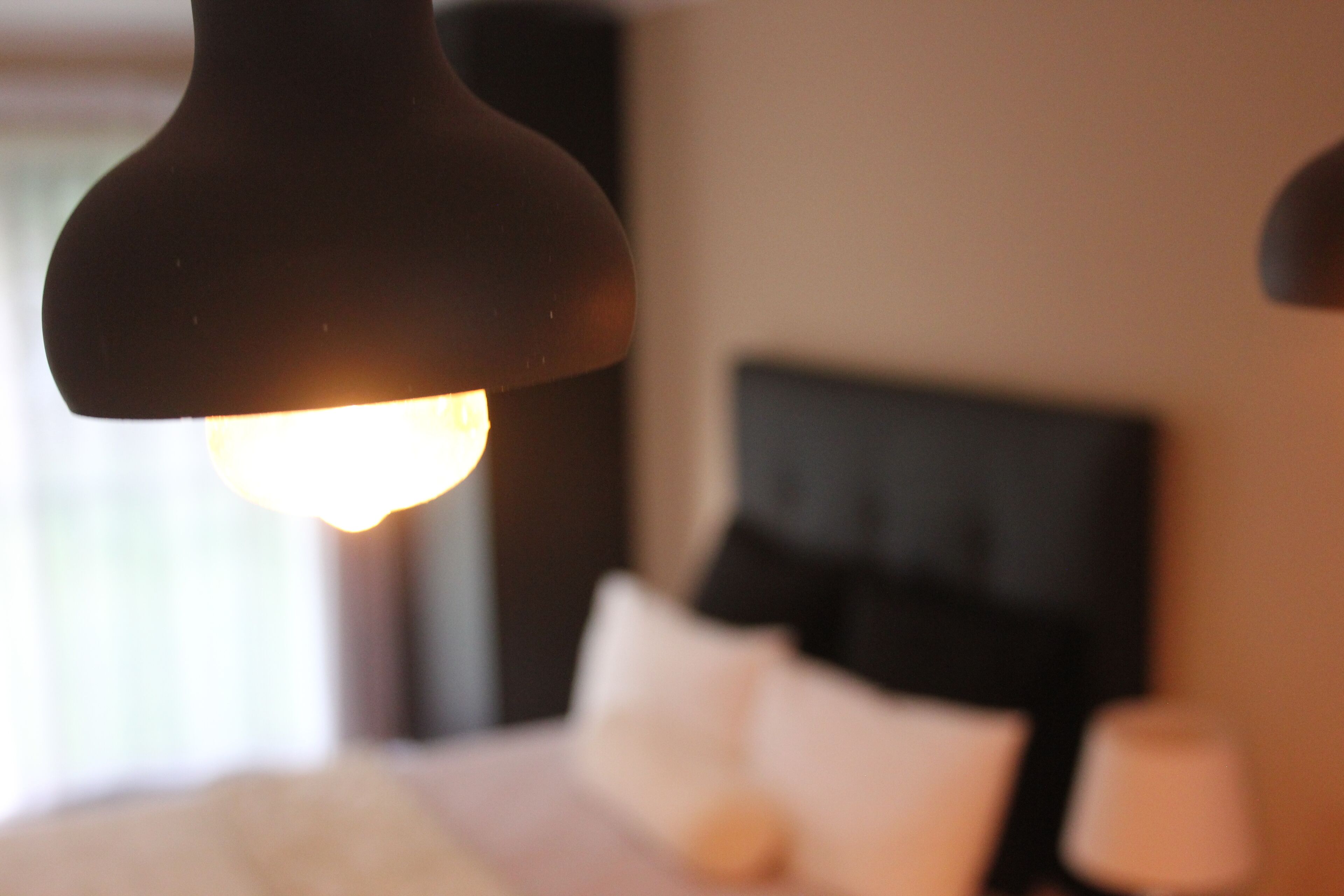 A close-up of a lamp with a warm light, a bed with white bedding, and a dark headboard in a hotel room.