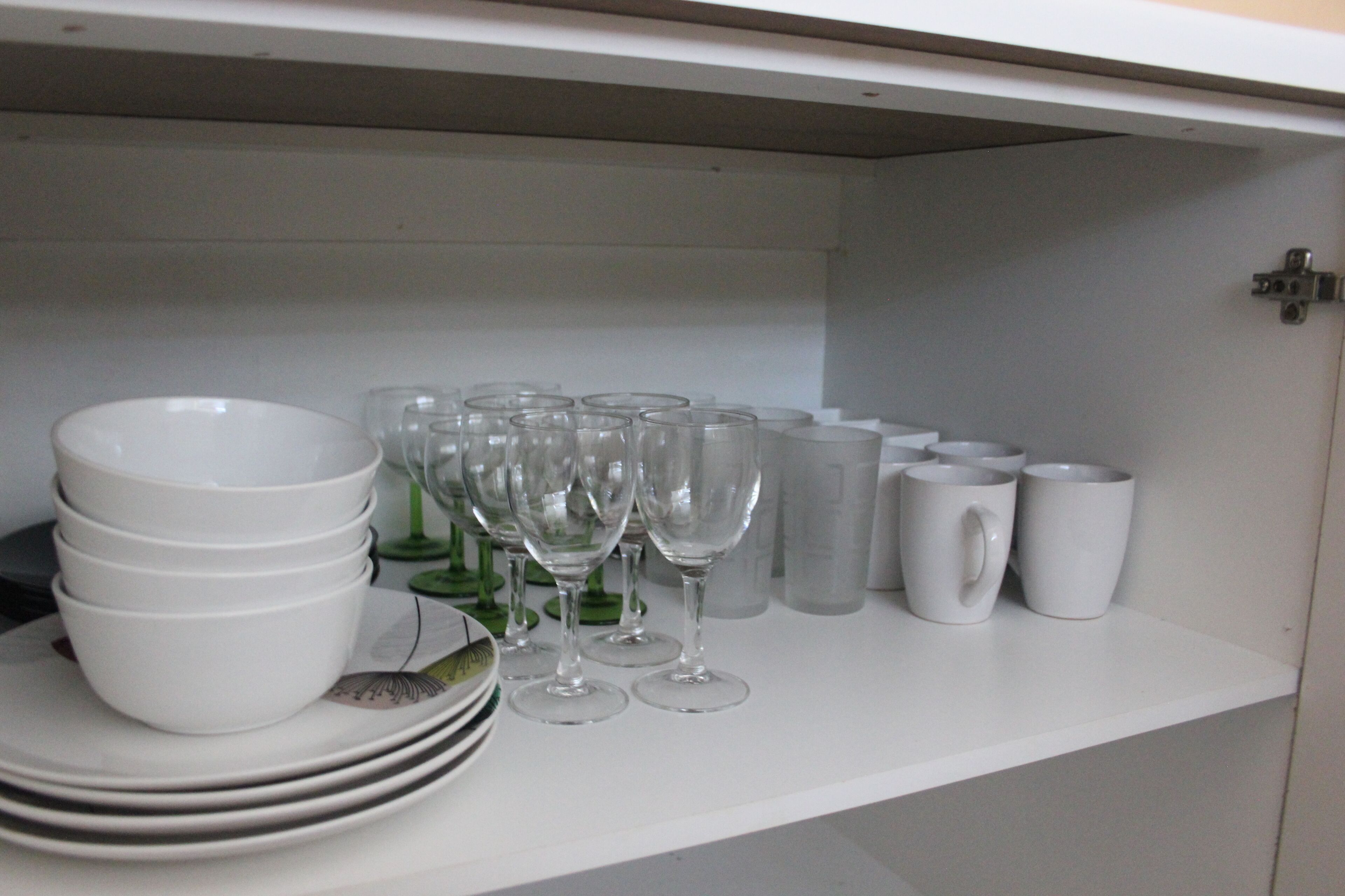 Un armario de cocina con cuencos blancos apilados, copas de vino y tazas blancas.