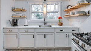A kitchen with white cabinets, a stove, a sink, and shelves with dishes and a coffee maker. - Charming Virginia Retreat w/ Covered Porch & Yard! (Farmville)