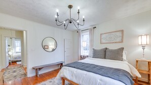 A bedroom with a large bed, wooden floor, a chandelier, a bedside table, a bench, and a mirror. - Charming Virginia Retreat w/ Covered Porch & Yard! (Farmville)