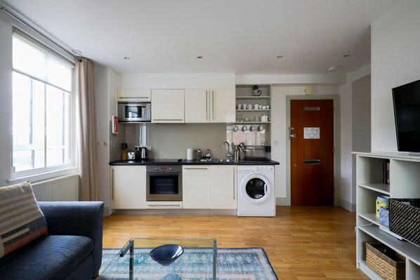 A compact kitchen with a microwave, oven, and washing machine, a sofa, and a TV on a stand. - Chelsea Pied-a-terre In Luxury Building (London)