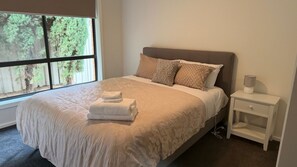 Een keurig opgemaakt bed met een beige dekbed en kussens, een nachtkastje met een lamp, en een raam met uitzicht op bomen.