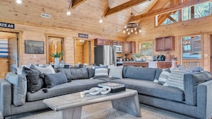 Interior moderno de cabaña de troncos con un amplio sofá seccional gris en forma de L, una mesa de comedor y una cocina con armarios de madera y un refrigerador.