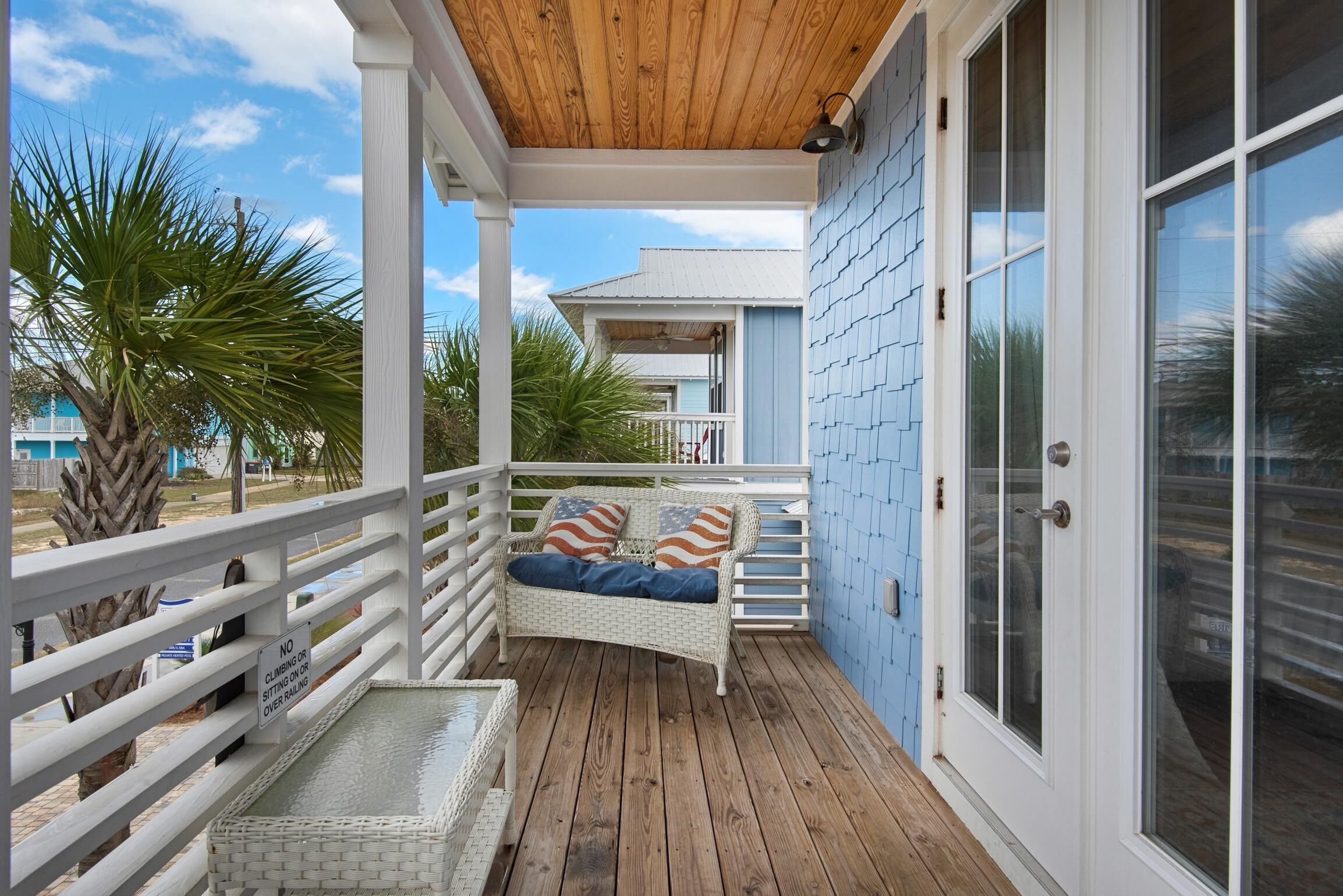 Eine überdachte Veranda mit weißer Geländer, ein Rattansofa mit Kissen mit amerikanischer Flagge und Blick auf ein blaues Gebäude im Hintergrund.