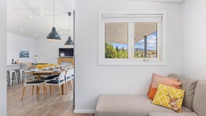 A modern living room with a sofa, a dining area with a table and chairs, a kitchen in the background, and a large window with a view of greenery.