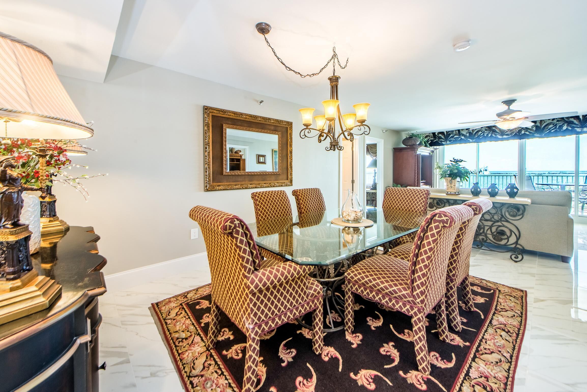 A dining area with a glass table surrounded by patterned chairs, a chandelier, and a large mirror on the wall.