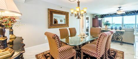 A dining area with a glass table surrounded by patterned chairs, a chandelier, and a large mirror on the wall.