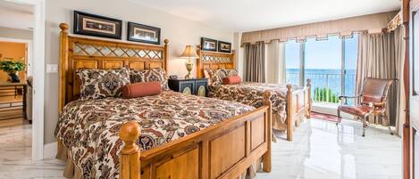 A spacious bedroom with a large bed, a nightstand, a lamp, and a view of the sea.