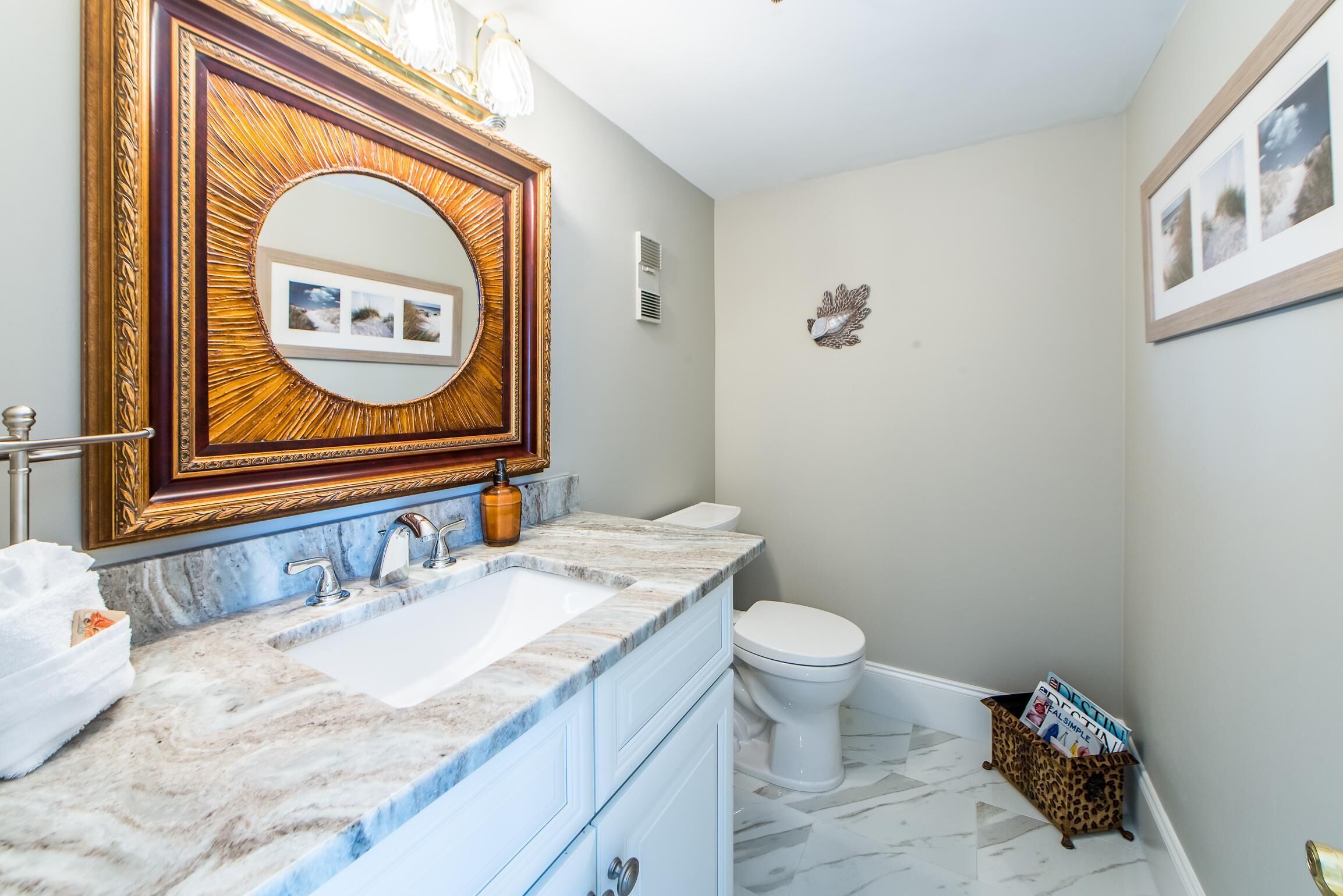 Une salle de bain avec un lavabo en marbre, un grand miroir orné d’un cadre décoratif, des toilettes et une petite étagère avec des magazines.