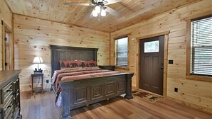 Un dormitorio de cabaña de madera con cama, mesita de noche, lámpara, ventilador de techo y ventana con persianas.