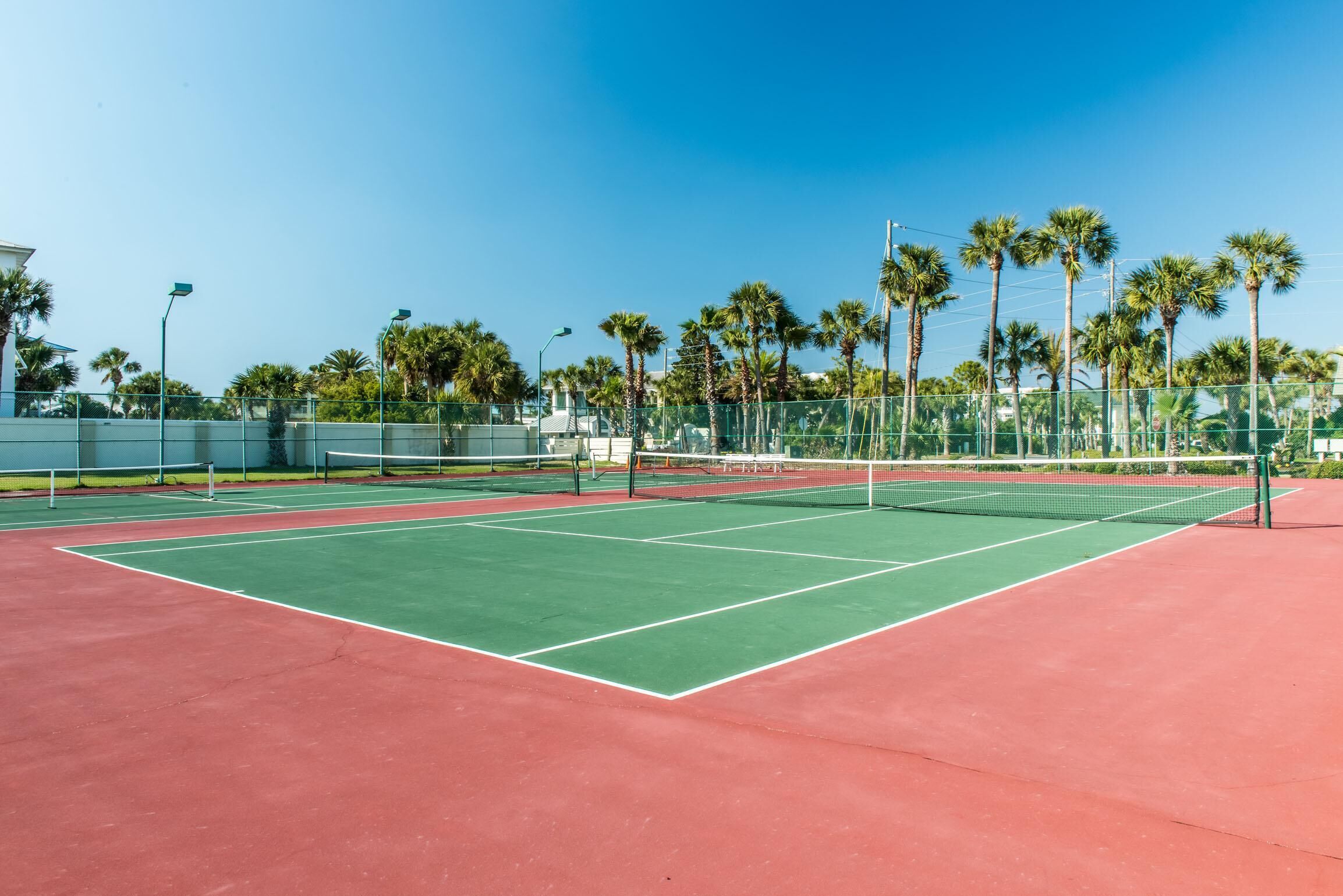 Un court de tennis doté d’une surface de jeu verte et d’une zone périphérique rouge, entouré d’une clôture métallique et de palmiers.