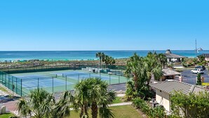 Un court de tennis offrant une vue sur l’océan et la plage, entouré de palmiers et d’un petit bâtiment.