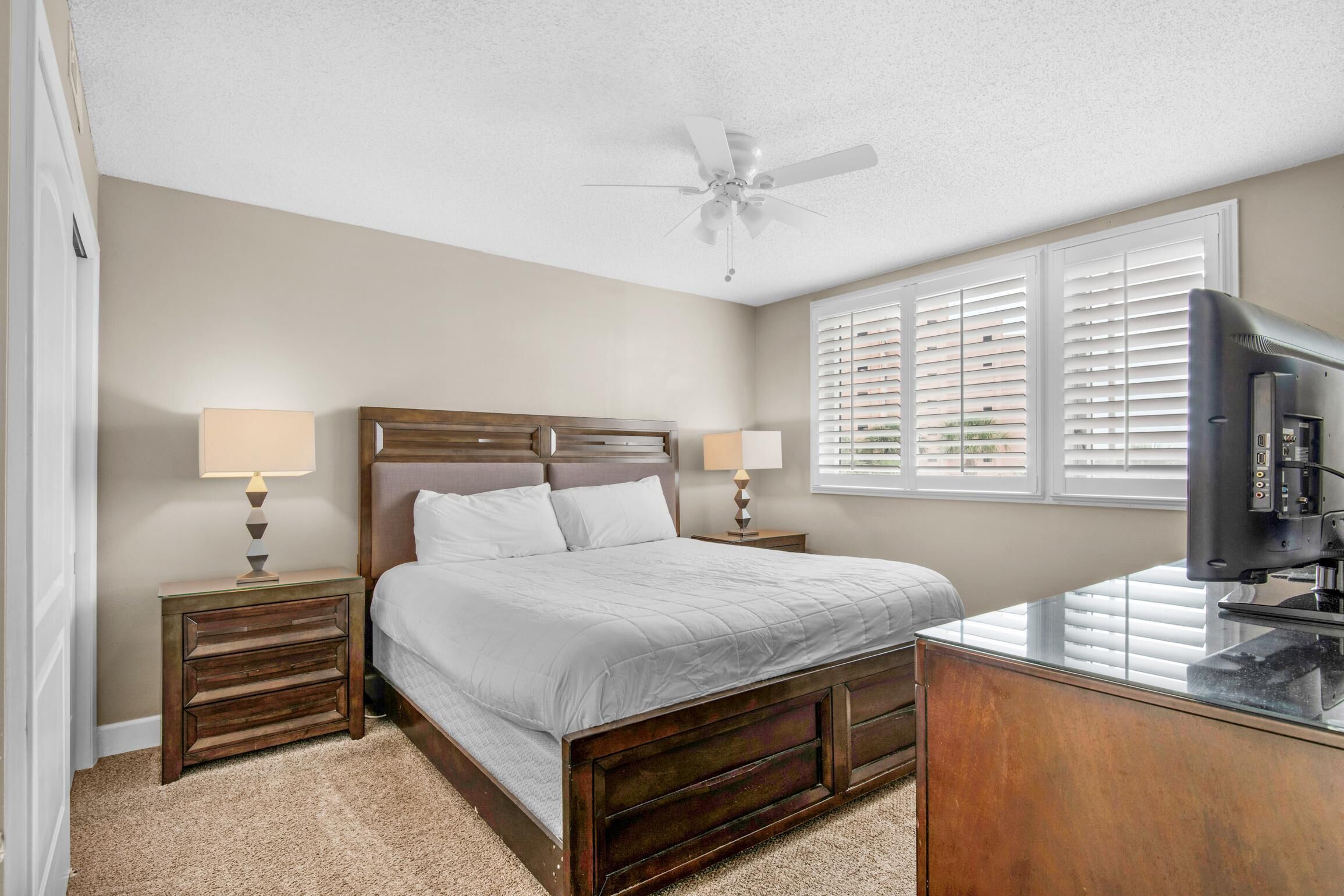 A bedroom with a bed, two bedside tables with lamps, a ceiling fan, a TV, and a window with blinds.