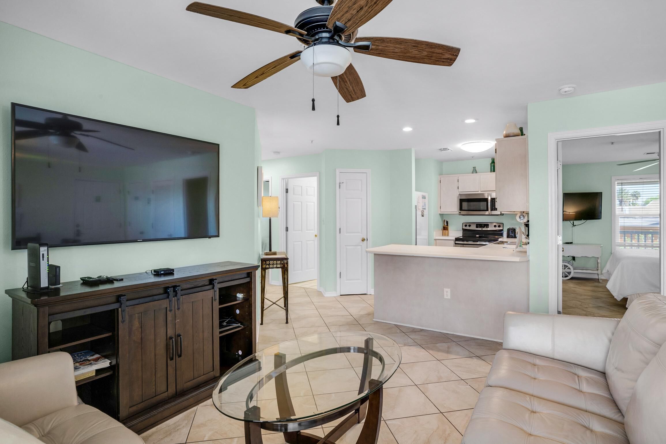 A spacious living room with a large TV, a glass coffee table, and a ceiling fan.