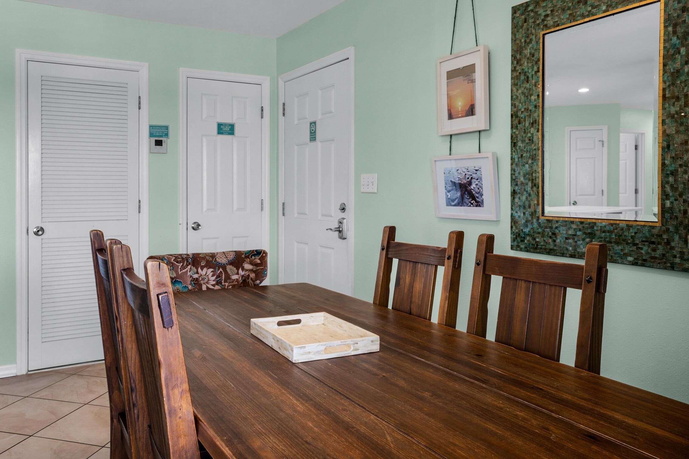 A dining area with a wooden table, chairs, and a bench. There are three doors, one with a sign, and a mirror on the wall.