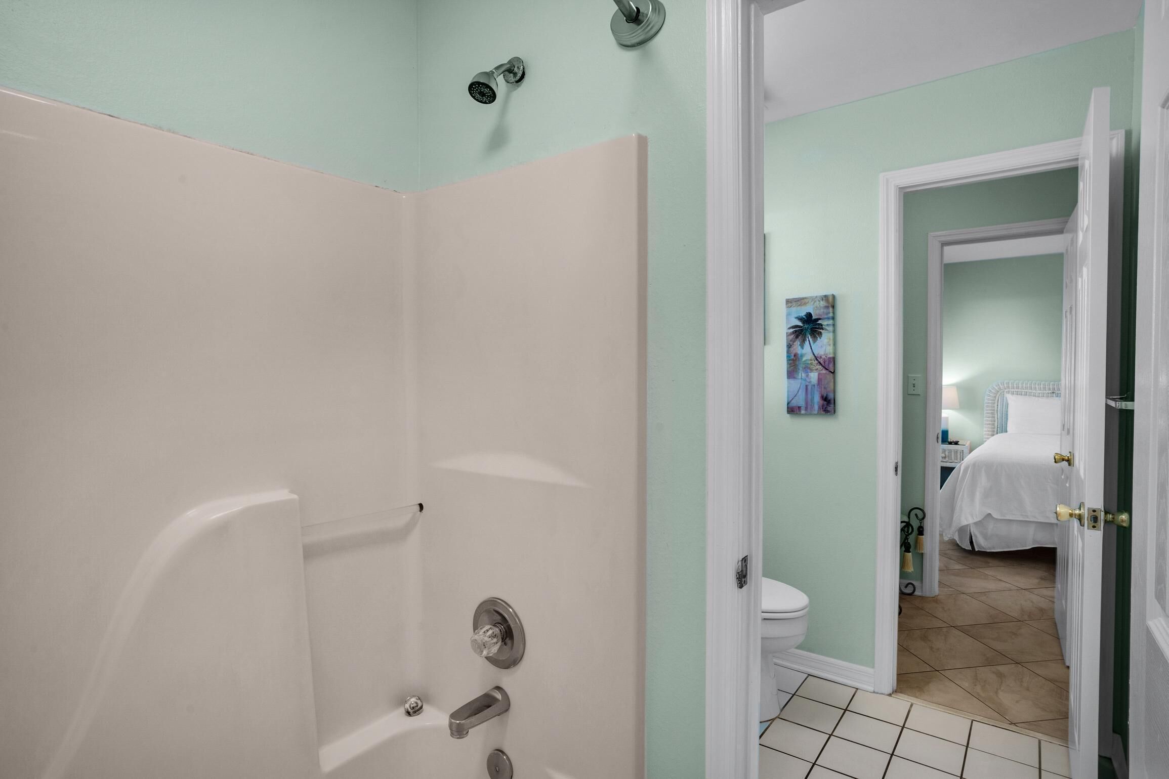 A bathroom with a shower, toilet, and a door leading to a bedroom with a bed and a nightstand.