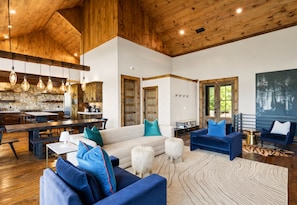 A spacious living area with a high ceiling, wooden beams, a stone fireplace, and a large window.