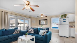 A modern living room with a blue sofa, white coffee table, ceiling fans, and a kitchen area with a refrigerator and stove. - St. Martin Beachwalk Villas 131 (Destin)
