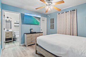 A bedroom with a bed, a ceiling fan, a TV, a bathroom with a toilet, and a shower curtain. - St Martin Beachwalk Villas 112 (Destin)