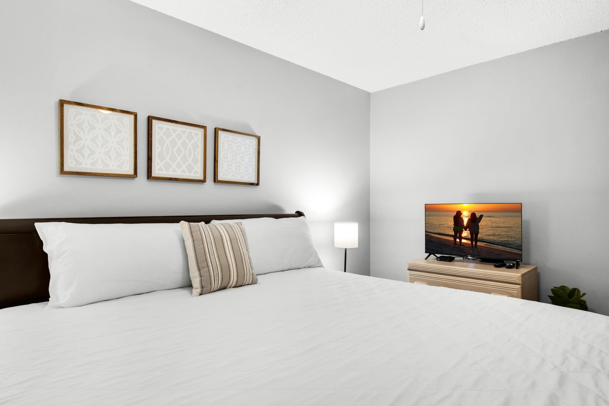 Une chambre d’hôtel avec un grand lit, un téléviseur diffusant une scène de coucher de soleil et trois tableaux encadrés accrochés au mur.