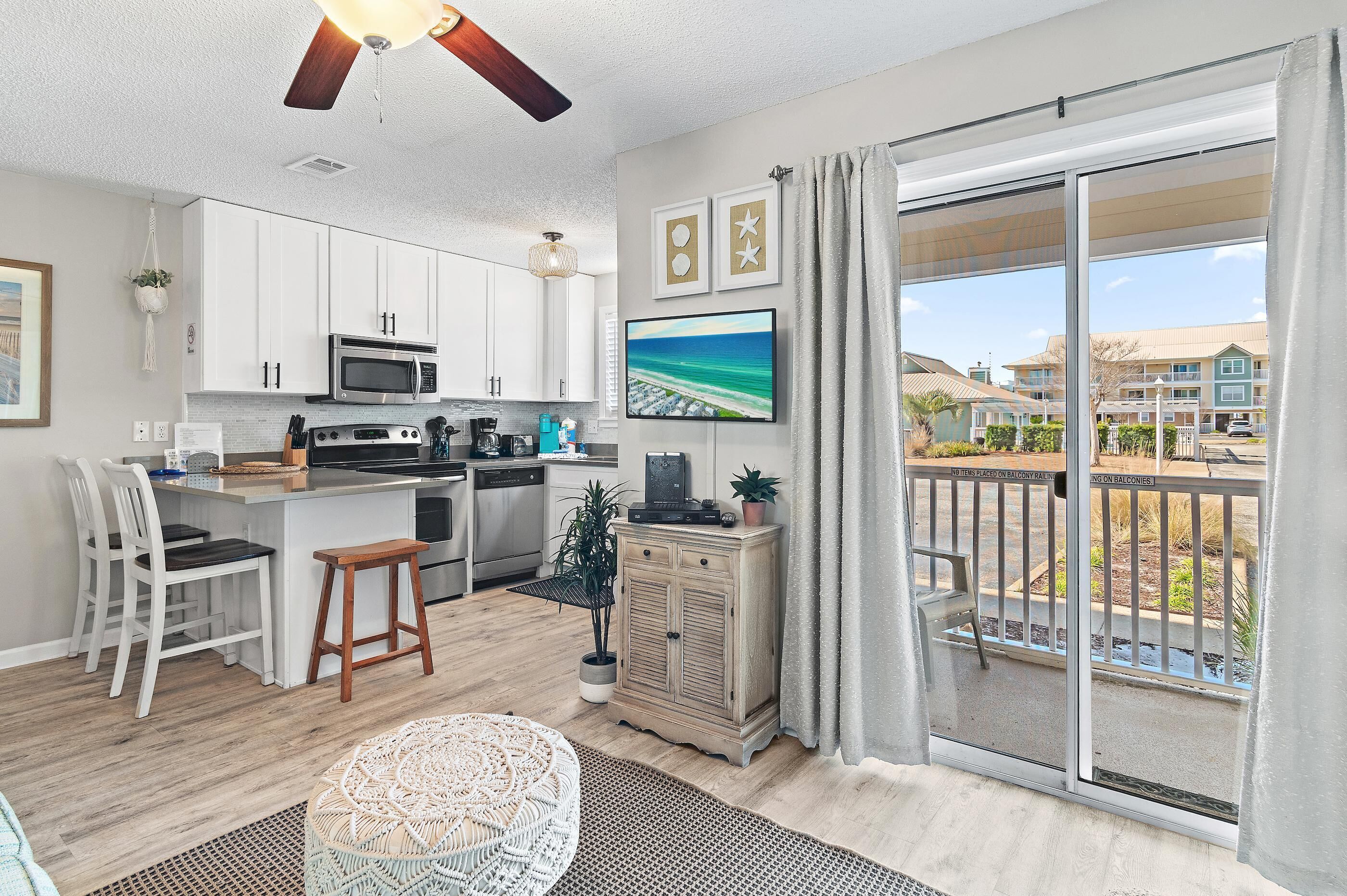 A modern kitchen with white cabinets, a microwave, and a large island. There is a dining area with white chairs and a wicker table. A sliding glass door leads to a balcony with a view of houses.