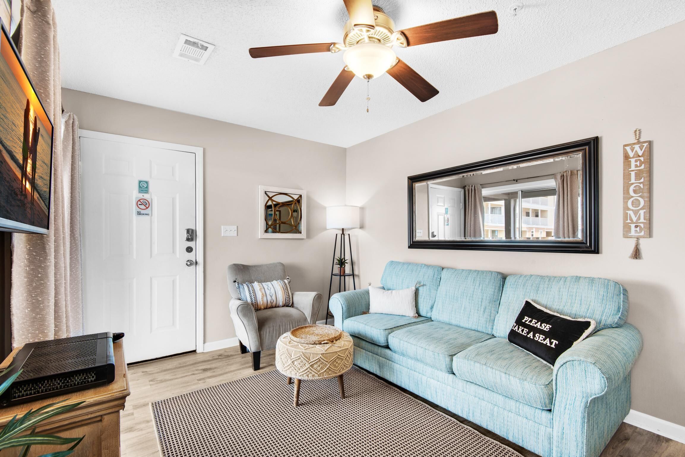 A living room with a ceiling fan, a light fixture, a sofa, a chair, a small ottoman, a rug, a framed mirror, and a welcome sign.
