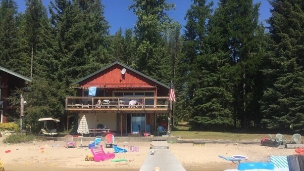 Une jetée en bois mène à un chalet doté d’un balcon, lequel est entouré d’une plage de sable et d’une forêt.