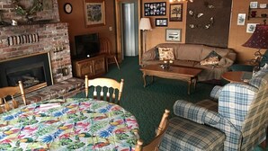 Un salon chaleureux avec une cheminée, un téléviseur, un canapé et une table à manger recouverte d’une nappe à motifs floraux.