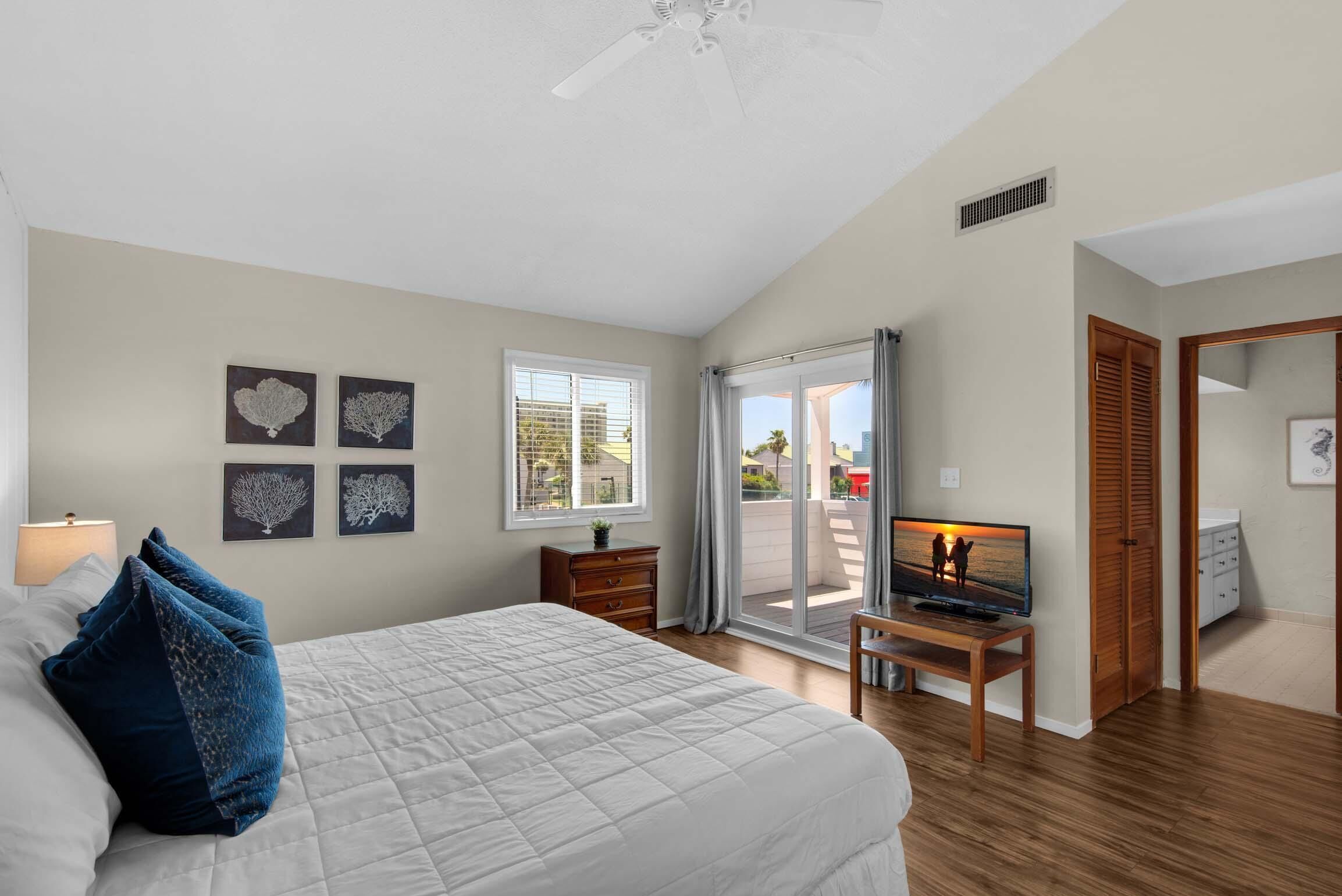 A bedroom with a bed, a TV on a wooden stand, a window with blinds, and a door leading to an outdoor area.