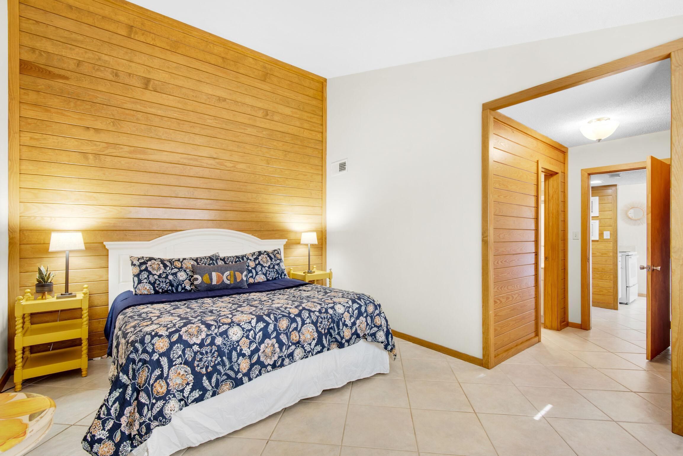 A bedroom with a wooden headboard, a floral bedspread, a bedside table with a lamp, and a wooden door leading to another room.
