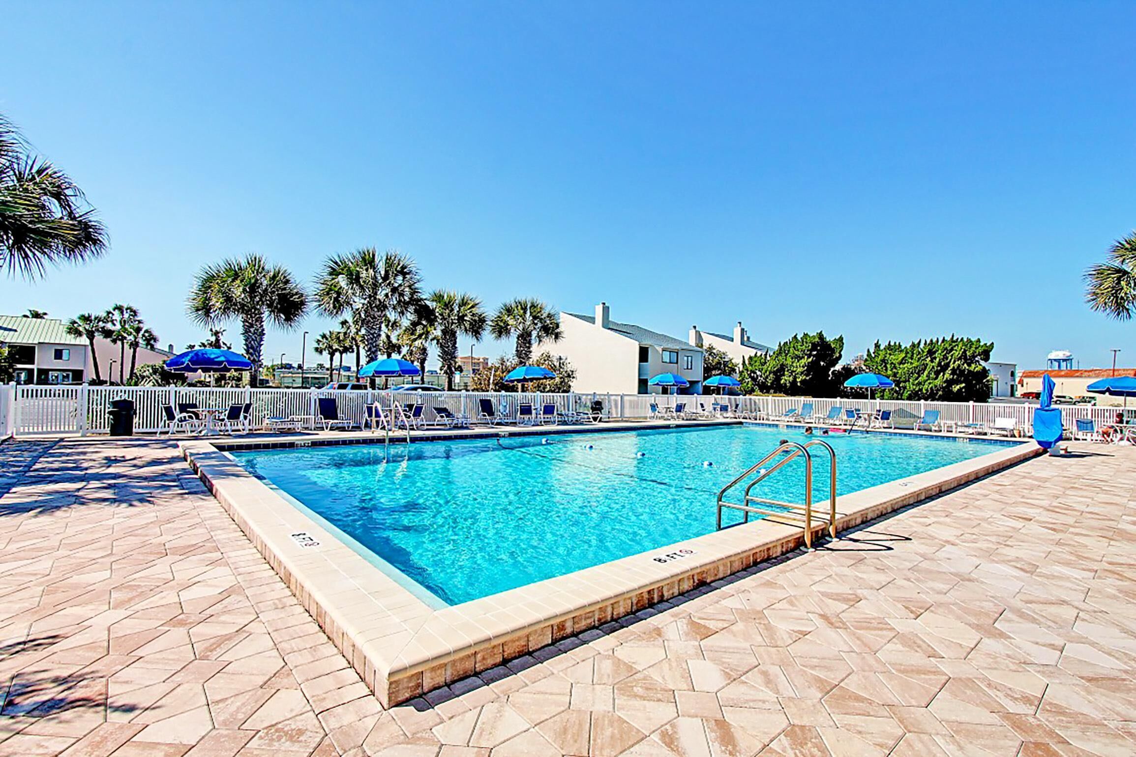 Ein Swimmingpool mit Fliesen umrandet, umgeben von Liegestühlen und Sonnenschirmen, mit Palmen und Gebäuden im Hintergrund.