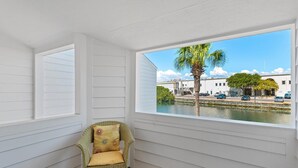 A room with a view of a water body and palm tree, a wicker chair with a cushion, and white paneled walls. - Shoreline Towers Townhouse 4-3 (Destin)
