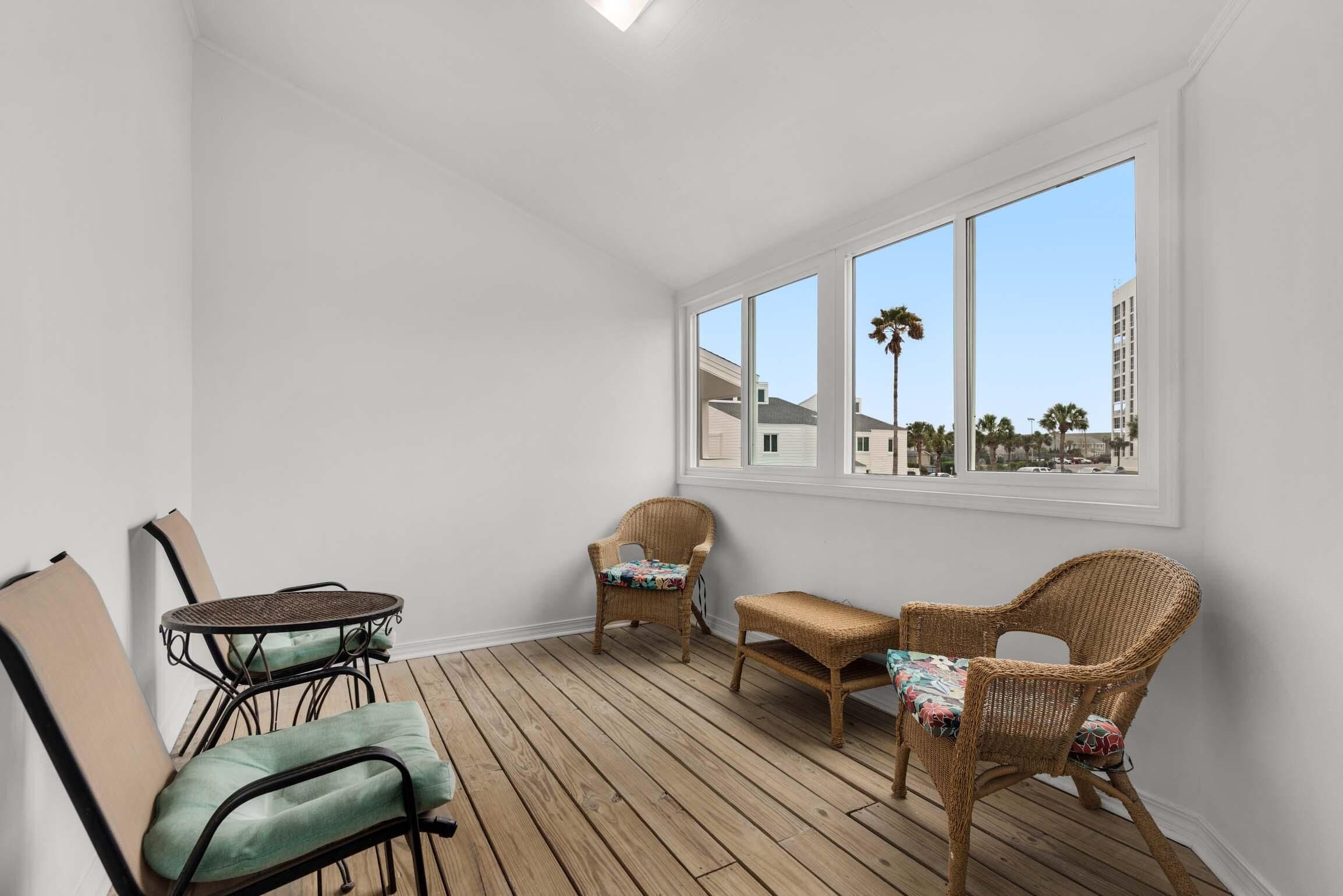 A sunroom with a wooden floor, white walls, and large windows offering a view of buildings and palm trees.