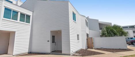 A modern two-story residential building with a white exterior, a front entrance, and a small front yard with mulch and a wooden fence.