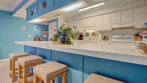 Een keuken met een wit aanrecht, blauwe wanden en witte kastjes. Er staan drie rieten krukken met beklede zittingen.