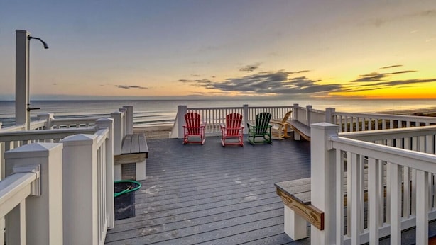 Een houten terras met witte leuningen, twee Adirondack-stoelen en uitzicht op de oceaan bij zonsondergang.