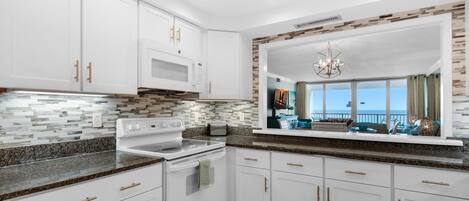A modern kitchen with white cabinets, granite countertops, a built-in microwave, and a large window overlooking a balcony.
