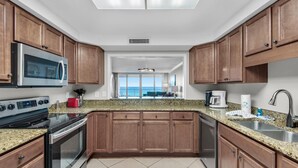 A modern kitchen with wooden cabinets, stainless steel appliances, and a large window offering a view of the outdoors. - Shoreline Towers 3092 (Destin)