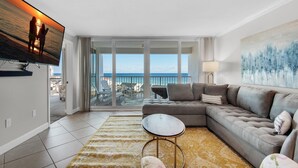 A modern living room with a large flat-screen TV, a grey sectional sofa, a small round coffee table, and a yellow patterned rug. - Shoreline Towers 3092 (Destin)