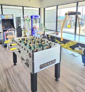 Een tafelvoetbaltafel met de aanduiding 'TORNADO' in een ruimte met grote ramen, speelautomaten en een stoel.