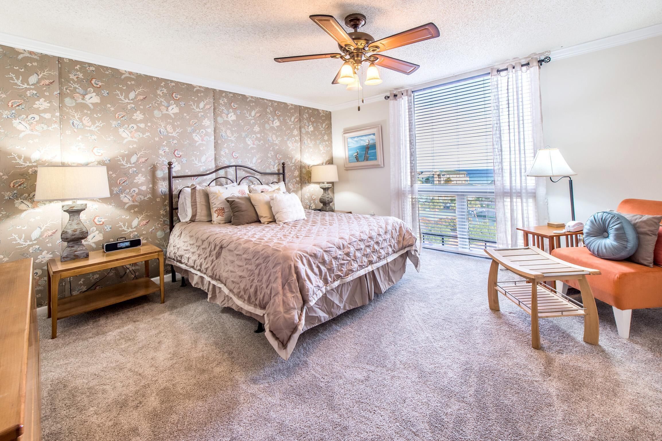 A bedroom with a bed, bedside tables, a ceiling fan, a window with blinds, and a view of the outside.