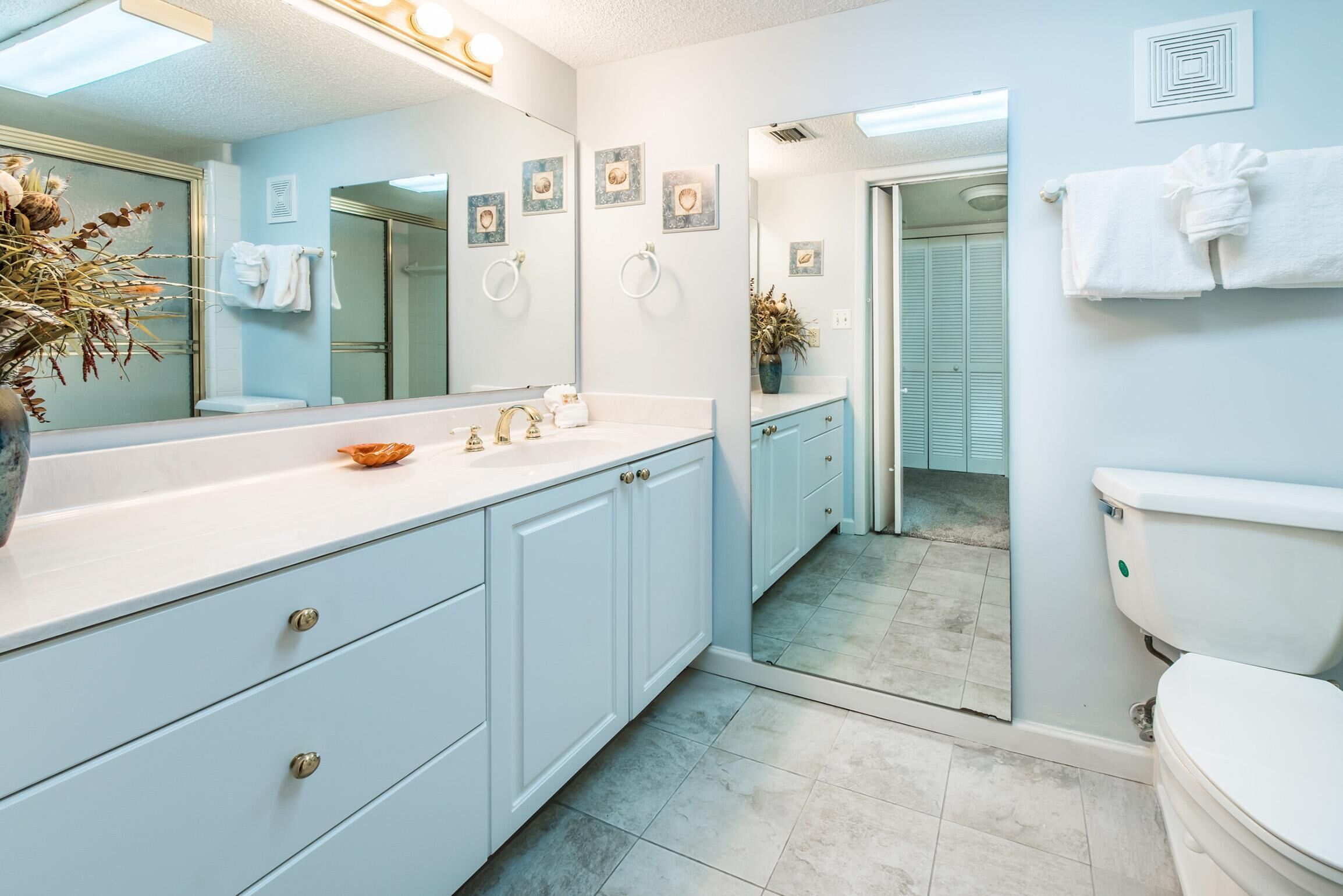 A modern bathroom with a large mirror, white sink, and light blue cabinets. There are decorative wall art pieces and a potted plant on the vanity.